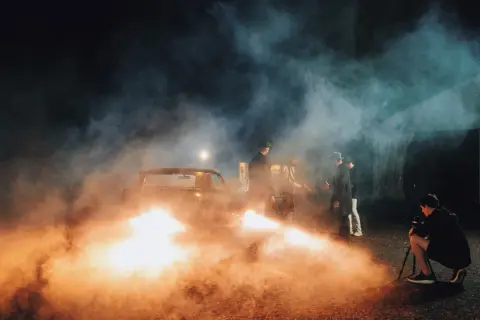 A film crew uses artificial light examples, including a bright spotlight and car headlights, to create a smoky, dramatic scene with men in hats gathered around a vintage car.