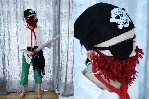 Dziecko w stroju pirata 122: czarna bandama z czaszką, opaska na oko, czerwona broda z włóczki, biała koszula, zielone spodnie i miecz.