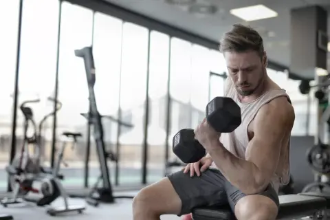 Mężczyzna ćwiczy biceps z hantlem, pokazując mięśnie synergistyczne przykłady. W tle siłownia.
