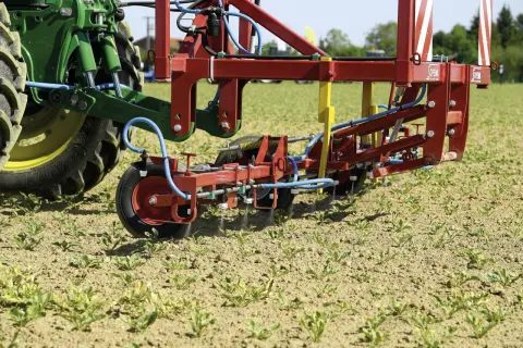Un tracteur vert et un pulvérisateur rouge travaillent dans un champ de jeunes pousses. La machine applique un traitement ciblé, peut-être pour une liste de graminées à éradiquer.