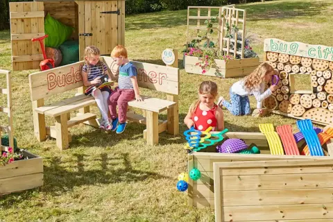 Children explore a vibrant Montessori garden, discovering nature's wonders. A bug city invites curiosity, while a planter box bursts with colorful blooms.