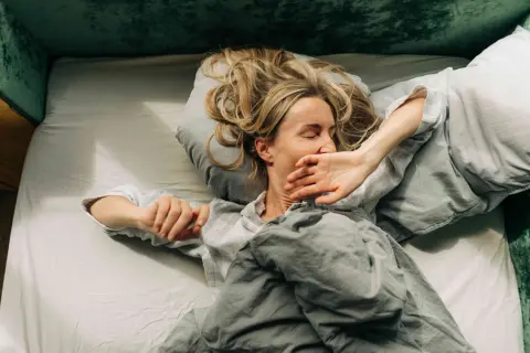 Woman in bed, covering her mouth, looking exhausted. Could this be narcolepsy vs hypersomnia?