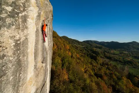 Ein Kletterer in orangefarbenem Shirt und roten Hosen erklimmt eine Felswand in Kroatien, während sich im Hintergrund ein herbstliches Tal erstreckt.