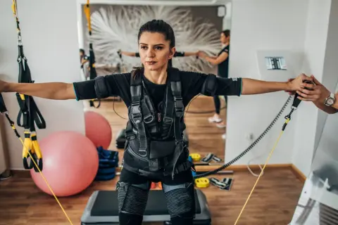 Kobieta w kamizelce do treningu EMS wykonuje ćwiczenia z taśmami. W tle widać drugą kobietę i sprzęt do ćwiczeń.