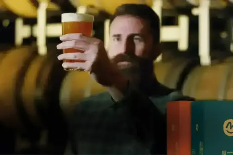 Uomo con barba esamina un bicchiere di birra dorata, con botti di legno sullo sfondo. Scatole di birre lager famose in primo piano.