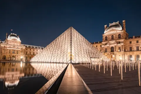 Das Louvre-Museum in Paris bei Nacht, mit der beleuchteten Glaspyramide im Vordergrund und dem historischen Gebäude im Hintergrund.