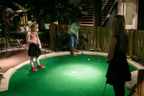 Barn spelar minigolf inomhus i Stockholm. En tropisk miljö med bambustaket och palmer.