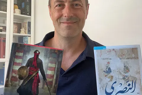 Juan de Granada, sonriente, sostiene dos libros. Uno muestra un guerrero con armadura y el otro, una escena histórica.
