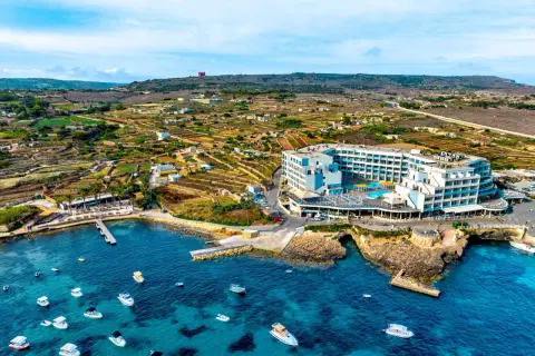 Malta last minute tanio: hotel z basenem nad turkusowym morzem, otoczony tarasami i łodziami. W tle malownicze, tarasowe pola i wzgórza.