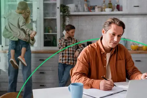 Mężczyzna w pomarańczowej koszuli pracuje przy laptopie, podczas gdy rodzina w tle cieszy się wspólnym czasem. Orange prąd w domu.