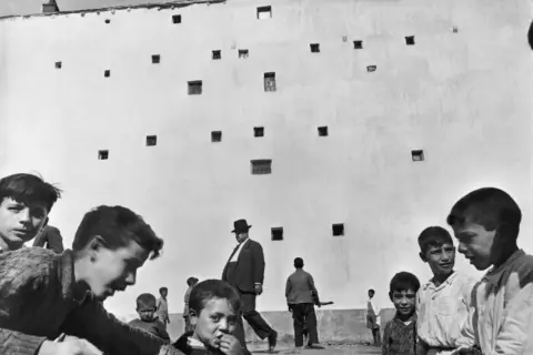 Enfants jouant devant un mur criblé de fenêtres, un homme en chapeau traverse cette photo historique célèbre.