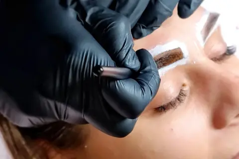 Stylist w czarnych rękawiczkach aplikuje hennę pudrową na brwi klientki.