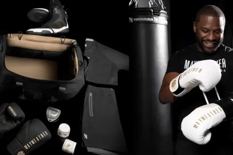 Floyd Mayweather Jr. smiles, holding a pair of white and gold Mayweather boxing gloves. The image also displays his boxing gear, including black gloves, shoes, and apparel.