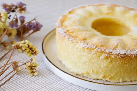 Delizioso chiffon cake ricetta originale americana, leggero e soffice, spolverato di zucchero a velo, con fiori secchi sullo sfondo.