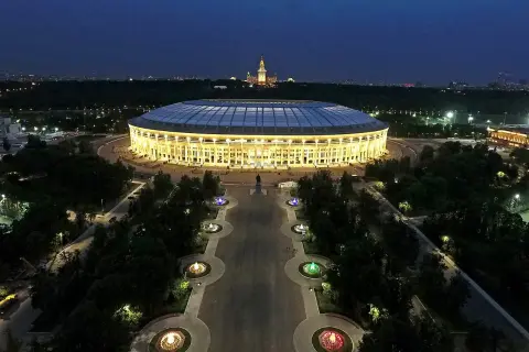 Oświetlony moskiewski stadion nocą, z aleją fontann i pomnikiem w tle.