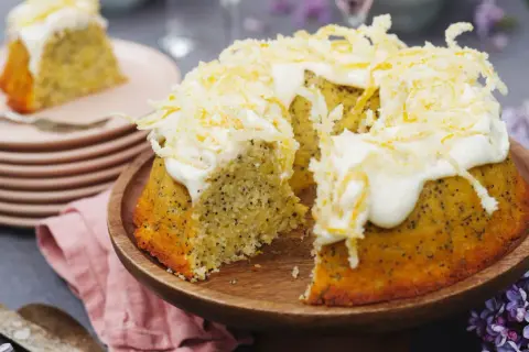 En saftig citron och vallmokaka med fluffig glasyr och citronskal. En bit är upplagd på en tallrik bredvid.