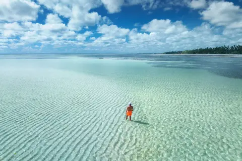 Człowiek w pomarańczowych spodenkach stoi w płytkiej, turkusowej wodzie. Zastanawia się, ile się leci na Zanzibar, by dotrzeć do tego raju.