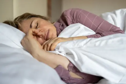 En kvinna sover 6 timmar i en säng med vita lakan och en lila rutig pyjamas. Hon håller handen mot ansiktet.