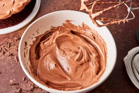 Schoko-Buttercreme in einer Schüssel, bereit zum Verzieren eines Kuchens. Ein Handmixer mit Schoko-Buttercreme hängt darüber.