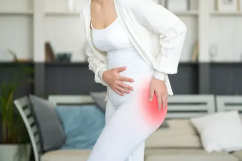 A woman in white clothing holds her hip, which is highlighted in red, indicating pain. She might be wondering, "why do my hips hurt when I sleep?