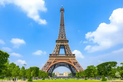 The Eiffel Tower stands tall against a bright blue sky with fluffy white clouds, surrounded by lush green trees and a manicured lawn.
