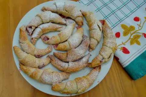 Pyszne, domowe rogaliki z marmoladą, posypane cukrem pudrem, ułożone na talerzu obok ręcznika z truskawkami.