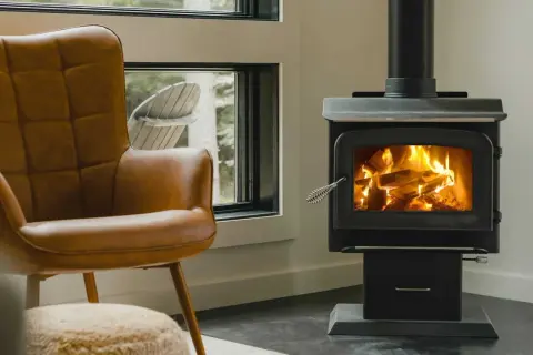 Un confortable fauteuil en cuir brun fait face à un poêle sans tubage crépitant, invitant à la détente près de la fenêtre.