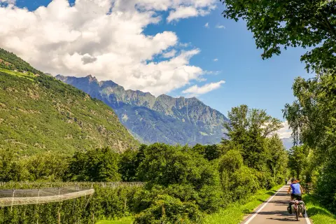 Cycliste sur la Via Claudia Augusta, traversant une vallée verdoyante bordée de montagnes majestueuses.
