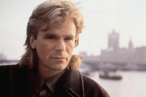 MacGyver obsada: Richard Dean Anderson w płaszczu, z charakterystyczną fryzurą, na tle zamglonego miasta.