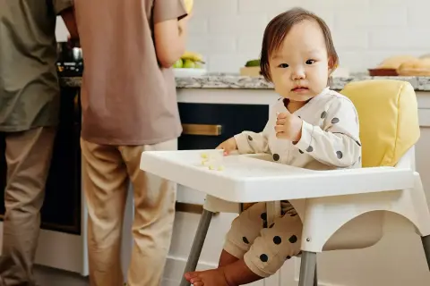 Małe dziecko siedzi w krzesełku dla dziecka, jedząc kawałki banana. W tle widać dwie osoby przygotowujące posiłek w kuchni.