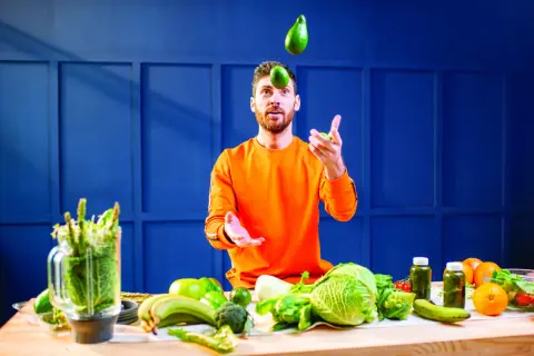 Un homme jongle avec des avocats, entouré de légumes frais. Une table garnie de fruits et légumes suggère une alimentation saine pour gérer l'acide urique.