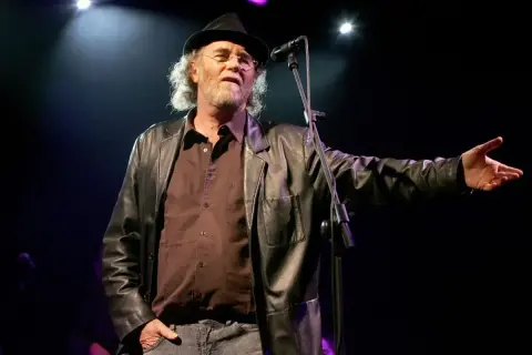 Francesco De Gregori, con cappello e giacca di pelle, canta sul palco. I suoi testi, come "Adelante Adelante", risuonano nell'aria.