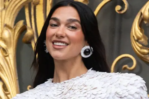 Uśmiechnięta Dua Lipa w białej kreacji, z dużymi kolczykami. Jej albańskie pochodzenie dodaje jej uroku.