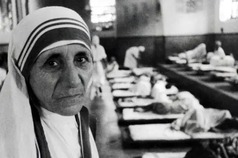 Mutter Teresa, deren Herkunft in Skopje liegt, blickt nachdenklich in die Kamera. Im Hintergrund liegen Kranke in Betten.