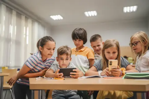 Grupa małoletnich uczniów skupiona wokół telefonu i tabletu, z zaciekawieniem oglądają coś na ekranach.