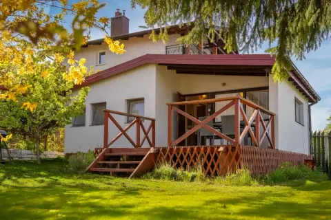 Bungalow z drewnianym tarasem i schodkami, otoczony zielonym ogrodem i kwitnącym drzewem.