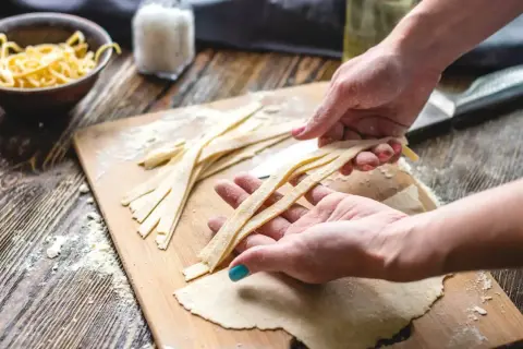 Hände formen frische Pasta-Streifen auf einem Holzbrett mit Mehl.