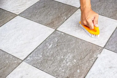 Main tenant, une brosse jaune frotte un carrelage blanc qui devient gris, révélant sa saleté.