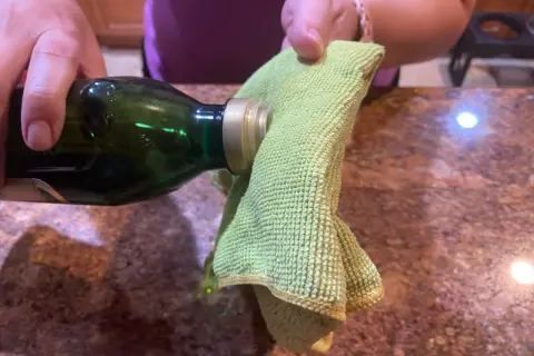A hand pours olive oil from a green bottle onto a green microfiber cloth, suggesting it's the best home remedy to clean stainless steel.