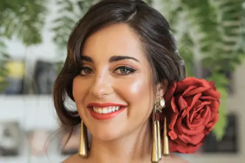 Peinados para feria pelo corto: mujer sonriente con rosa roja en el cabello, pendientes dorados y maquillaje festivo.
