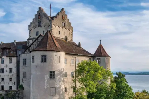 Das Meersburg Castle thront über dem Bodensee, umgeben von grünen Bäumen und unter einem blauen Himmel mit Wolken.
