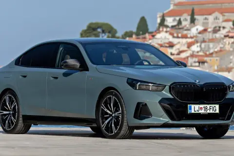 Szary sedan BMW G60 z czarnymi felgami i grillem, zaparkowany na tle nadmorskiego miasteczka.