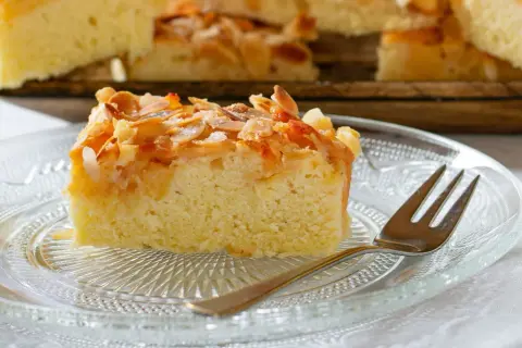 Ein saftiges Stück mandelkuchen vom blech, bestreut mit gehackten Mandeln und Zucker, auf einem Glas-Teller mit einer Gabel.