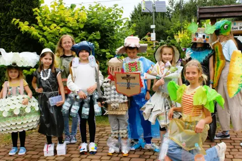 Dzieci w strojach z recyklingu, wykonanych z plastiku, papieru i kartonu, pozują do zdjęcia na zewnątrz.