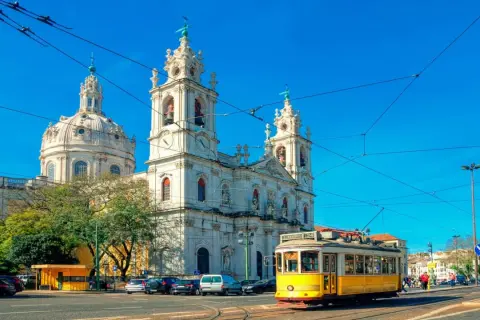 Un tranvía amarillo cruza frente a una iglesia blanca con cúpulas y torres, reflejando cómo se vive en Portugal con su encanto histórico y transporte icónico.
