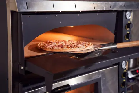 Une pizza garnie est en cours de cuisson dans un four professionnel, sur une pelle à pizza. La chaleur intense du four promet une cuisson parfaite.