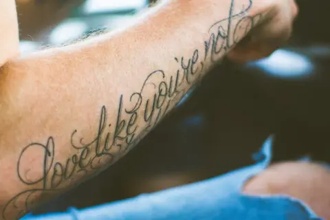Ein Arm mit einem geschwungenen Schriften Tattoo, das die Worte "Love like you're not" trägt.