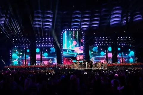 Wydarzenia na Stadionie Narodowym: scena z neonowymi samochodami i tańczącymi artystami, oświetlona przez reflektory. Tłum widzów wpatrzony w widowisko.