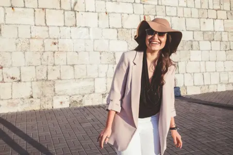 Donna sorridente con cappello e occhiali da sole, perfetta per come vestirsi con 20 gradi: blazer leggero, top nero e pantaloni bianchi.