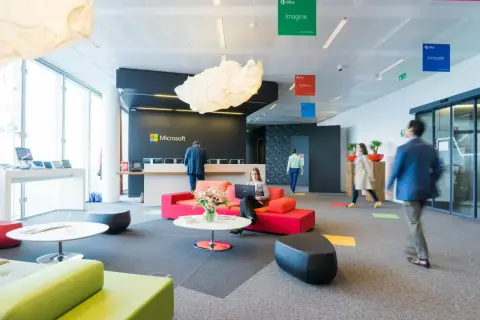 Un **open space** moderne chez Microsoft. Une femme travaille sur son ordinateur portable, entourée de canapés colorés. Des affiches "Imagine", "Achieve", "Innovate" flottent au plafond.